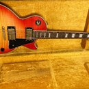 Elektrická kytara s červeně žlutým gradientem (sunburst) a viditnou strukturou dřeva v hnědém tvrdém kufru. Na hlavě je bílé logo s nápisem Epiphone.…