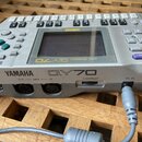 Stříbrný sekvencer s displejem a množstvím tlačítek. Na přední straně je nápis YAMAHA QY70 MUSIC SEQUENCER a v levém rohu loga XG a MIDI. Zadní panel…