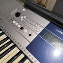 Detailní pohled na ovládací panel a klávesy elektronického klavíru v modré a stříbrné barvě. Na pravé straně je viditelné logo YAMAHA a malý displej.…