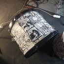 Fotografie zobrazuje basový buben s černými obručemi. Korpus je opatřen bílo-černým potiskem v komiksovém stylu s různými postavami a prvky. V pravé…