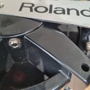Detailní záběr na černý plastový prvek s kovovým štítkem. Na štítku je černý nápis Roland a neúplný nápis PD-. Povrch plastu vykazuje drobné prachové…