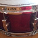 Fotografie znázorňuje snare drum s tmavě červeným dřevěným korpusem a chromovaným kováním. Na horním obruči je nápis SONOR 2.3mm PowerHoop doprovázen…