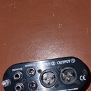 Černý oválný konektor s několika vstupy a výstupy. Na zařízení jsou viditelné nápisy OUTPUT 1 (u dvou konektorů), OUTPUT 2 (u dvou konektorů) a INPUT…