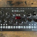 Černý syntezátor s nápisem BASSLINE a označením DB-01 v levém dolním rohu. V pravém dolním rohu je logo erica synths. Přístroj obsahuje řadu černých…