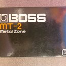 Černá krabice s bílým logem BOSS. Pod ním je oranžový nápis MT-2 a bílý text Metal Zone. V pravém dolním rohu je malý bílý štítek s nápisem USE BOSS…