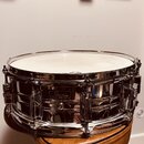 Stříbrný kovový malý buben s bílým koženem, umístěný na hnědém polstrovaném sedadle. Na korpusu je viditelné logo "Pearl". Povrch nástroje je vysoce…