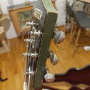 Detail hlavy kytary v olivově zelené barvě. Na čele je viditelný nápis GRETSCH. Jsou zde vidět tři chromované ladičky a struny; povrch lakování vykaz…