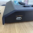 Detailní pohled na černý plastový přístroj s USB portem. Nad konektorem je žlutý nápis USB STORAGE. Na horní části jsou vidět černé ovládací tlačítka…