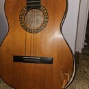 Kytara s přírodním dřevěným povrchem korpusu a černým hmatníkem. Uvnitř otvoru rezonanční desky je viditelný nápis „Lutona CLASSIC GUITAR“. V pravém…
