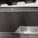 Detailní pohled na černý přístroj s texturovanou protiskluzovou plochou. V pravém dolním rohu je umístěno stříbrné logo s nápisem HEADRUSH. V horní č…