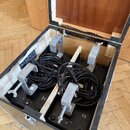 Otevřený transportní kufr z dřeva a překližky, v němž jsou uloženy dva svazky černých kabelů. Kabely jsou fixovány k černé základní desce pomocí čtyř…