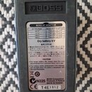 Zadní strana efektového pedálu značky BOSS v šedé barvě. Na spodní části je bílý štítek s tabulkou BOSS AC ADAPTOR, nápisem Dry battery 9 V a varovný…
