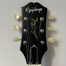 Černá hlava elektrické kytary s bílým nápisem Epiphone v horní části. Uprostřed je umístěn žlutý nápis Les Paul MODEL. Nástroj má šest ladících kolíč…