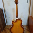 Fotografie zachycuje zadní stranu akustické kytary s vyřezem v těle. Nástroj má lakovaný povrch v přechodovém odstínu od žluté k tmavě hnědé (sunburs…