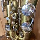 Detailní pohled na část saxofonu v mosazném provedení. Nástroj vykazuje známky opotřebení povrchu v podobě drobných škrábanců a oxidace. Na fotce nej…