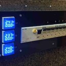 Černý rackový distribuční panel s třemi modrými digitálními voltmetry a ampérmetry označenými L1, L2 a L3. Uprostřed je umístěn bílý proudový jistič…