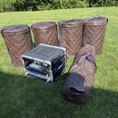 Na trávě stojí čtyři hnědé prošívané obaly na bubny a jeden podobný dlouhý obal na nástroj. Mezi nimi je umístěn černý transportní flightcase s kovým…