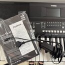 Na fotografii je černý digitální klavír značky YAMAHA. Na nástroji leží v plastovém obalu uživatelský manuál s nápisy YAMAHA, PSR-SX, Digital Worksta…