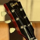 Fotografie zachycuje hlavu elektrické kytary s černým povrchem a červeným lemováním. Na čelní straně je zlatý nápis Gibson a pod ním kurzivou nápis L…