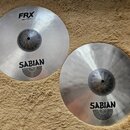 Dvě stříbrné činely značky Sabian s matným povrchem. Levý činel nese nápisy FRX, Hats 14"/35 cm a logo SABIAN. Pravý činel má pouze logo SABIAN. Oba…