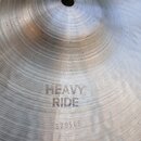 Detailní pohled na povrch kovového činelu s viditelným opracováním v podobě koncentrických rýh a kování. Na povrchu je vyražen nápis HEAVY RIDE a níž…