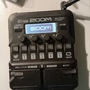 Černý multi-efektový procesor s nápisy G1 FOUR, ZOOM a Multi-Effects Processor. Zařízení má displej s logem ZOOM, čtyři otočné ovladače označené Lo,…