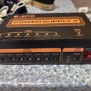 Černý napájecí adaptér s oranžovými prvky a nápisy. Na horní straně je logo JOYO a velký nápis POWER SUPPLY 4. Přední panel obsahuje sedm výstupů DC…