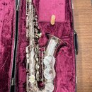 Stříbrný saxofon v černém kufru s fialovým polstrováním. Nástroj vykazuje známky opotřebení v podobě drobných škrábanců a znečištění povrchu. Na těle…