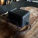 Černý síťový adaptér s hranatým tvarem a připojenými kabely. Na horní části je viditelný nápis AC-AC ADAPTOR. Povrch plastového krytu vykazuje drobné…