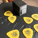 Na černém povrchu leží osm žlutých trsátel s nápisy „DUNLOP“, „TORTEX“, „U.S.A.“ a „73 mm“. Uprostřed je umístěn malý černý kostkový box s viditelnou…
