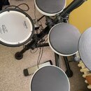 Elektronická bicí souprava s černým rámem a bílými okraji padů. Na jednom z padů je čitelné logo Roland a nápis No. 123456789 (nečitelné). Další pad…