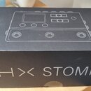 Černá papírová krabice s nápisem HX STOMP. Na horní části krabice je bílou čarou znázorněna schéma efektového pedálu. Povrch krabice vykazuje mírné p…