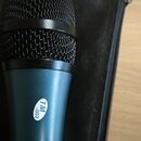 Detailní pohled na horní část mikrofonu s černou kovovou mřížkou a tmavě modrým tělem. Na těle je nalepená bílá oválná nálepka s nápisem TM -002. Mik…
