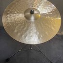 Zlatý kovový činel umístěný na černém stativu. Na povrchu je nápis „Paiste“ a označení „Dry Ride 20"“. Povrch činelu je zdobný koncentrickými rýhami…