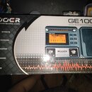Kartonová krabice s produktem MOOER GE100 Guitar Multi-Effects Processor. Na obalu je zobrazen přístroj s displejem, na kterém je nápis SCALE LESSON…