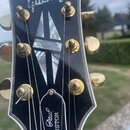 Detail hlavy černé elektrické kytary s bílým lemováním. V horní části je bílé logo Gibson a v dolní části nápis Les Paul CUSTOM. Uprostřed se nachází…