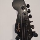 Fotografie zachycuje hlavu elektrické kytary v černé barvě. Na povrchu je zlatý nápis Squier a vedle něj menší bílý text BY FENDER® STAGEMASTER®. Nás…