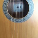 Detailní pohled na ozvukový otvor akustické kytary s dřevěnou deskou světlé barvy. Uvnitř otvoru je štítek s nápisy Cort GUITARS, MODEL: AC10 NS a SE…