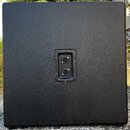 Zadní strana černého reproduktorového boxu s texturovaným povrchem. Uprostřed se nachází připojovací panel se dvěma vstupy typu jack. Povrch v dolní…