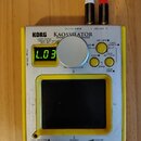 Stříbro-žlutý přenosný syntetizátor s nápisy KORG a KAOSSILATOR DYNAMIC SYNTHESIZER. Zařízení má malý digitální displej s nápisem L.03, jeden velký s…