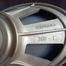 Detailní pohled na kovový reproduktor stříbrné barvy. Na povrchu je vyražen nápis GRUNDIG v oválném rámu a pod ním označení 360 - □. Povrch vykazuje…