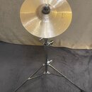 Zlatavý kovový činel umístěný na chromovaném stativu s třemi nohami. Na pravé straně činelu je viditelný nápis "Paiste". Povrch činelu vykazuje jemné…