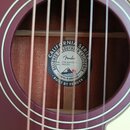 Detail zněku akustické kytary v tmavě červené barvě. Uvnitř zněku je vidět kulatá štítková nálepka s logem Fender a nápisy CALIFORNIA SERIES, DESIGNE…