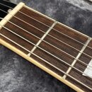 Detail krku elektrické kytary s hnedým hmatníkem a bílými bindingy. Na černé hlavě je viditelna část bílého loga ve tvaru stylizovaného písmene. Hmat…