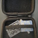 Otevřený černý zipový pouzdro s vnitřním polstrováním a síťovou kapsou. V kapse je umístěna kovová harmonika a papírový obal s modrým nápisem, ze kte…