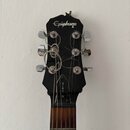 Fotografie zachycuje hlavu černé elektrické kytary s logem Epiphone. Na horní hraně hlavy je viditelné odloupaní laku a poškození materiálu. Mezi lad…