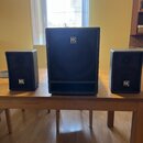 Tři černé reproduktory s mřížkami v provedení kosočtverců, umístěné na dřevěném stole. Uprostřed je jeden větší reproduktor a po stranách dva menší.…