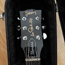Fotografie zachycuje hlavu elektrické kytary v černém kufru s plyšovou výstěvou. Na černé hlavě je zlatý nápis Gibson a bílé nápisy Les Paul 100 a St…