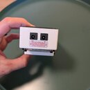 Fotografie zachycuje zadní stranu bílého efektpedálu s dvěma černými konektory pro napájení. Vedle konektorů jsou nápisy IN a OUT. Pod nimi je bílá n…