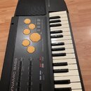 Černý elektronický klávesový nástroj s bílými a černými klávesami. Na levé straně je nápis DYNAMIC BASS a sekce s Percussion Pad tvořená šesti oranžo…